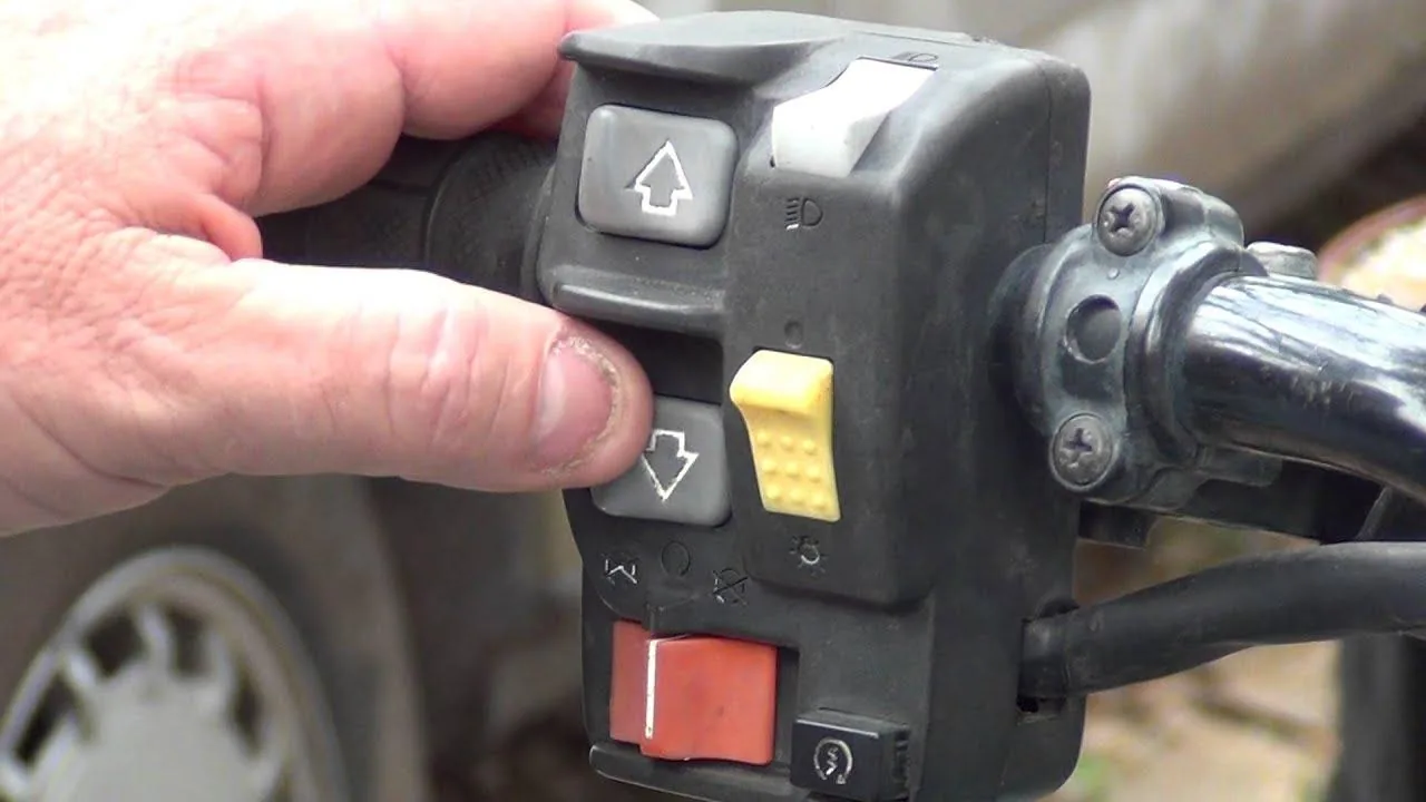 Close-up of a hand on the left handlebar switch of an older electric-shift Honda ATV, with up and down shift buttons